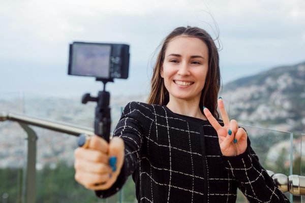 Smiling blogger girl is taking selfie by showing victory gesture against the background of city view.